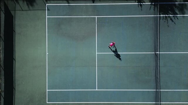 A female tennis player in a pink shirt practices on a hardcourt, showcasing agility and focus. Drone footage captures her dynamic movements, highlighting skill development and intense training.