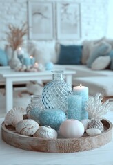 A light-filled living room scene displays a decorative tray filled with seashell accents, teal-toned glass items, and candles in a serene coastal setting.