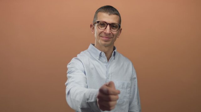 Man wearing glasses extends hand for handshake against brown wall, exuding friendly and approachable demeanor in isolated setting with focus on interpersonal connection.
