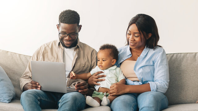 Happy Loving Family Of Three Using Laptop Sitting On Couch After Moving New Home. Real Estate, House Ownership And Relocation Concept. Afro People Browsing Internet Sitting Among Carton Boxes