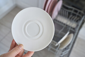 Hand holding clean white saucer from dishwasher in modern kitchen setting.