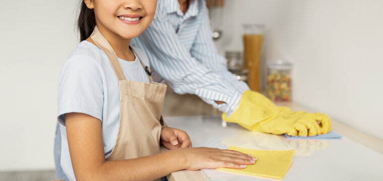 Young asian mother teaching her daughter housekeeping, cleaning kitchen together and smiling at camera, selective focus. Cute girl wiping table with cloth, enjoying helping with household chores