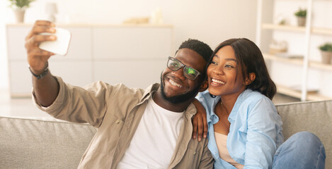 Portrait of happy African American couple taking selfie together at home indoors. Excited lady and guy in braces posing and holding smartphone, sitting on the couch in living room, looking at screen