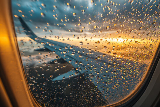 View from airplane window covered with rain droplets showing wing during vibrant orange and blue cloudy sunset sky with soft light reflections on the glass - Powered by Adobe