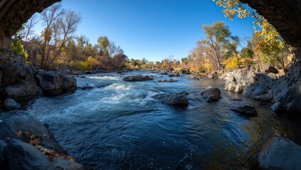 A serene river scene, flowing swiftly amidst rocks and trees under a clear, bright sky, framed by a natural archway.