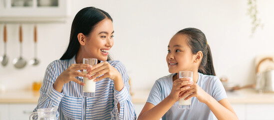 Pretty asian teen girl drinking milk with her mom and eating cookies, sitting at kitchen table,...