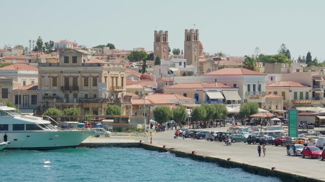 Mediterranean coastal town. Marina with docked yacht, waterfront promenade, red-roof buildings, and twin church towers in a busy seaside town.