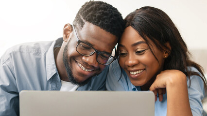 Portrait of happy loving young African American couple using laptop, watching video, looking at their photos or choosing what to buy, sitting and relaxing on floor in living room