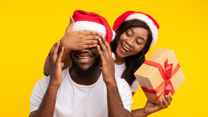 Merry Christmas. Black Girlfriend Surprising Boyfriend With Xmas Gift Covering His Eyes Posing Wearing Santa Hats Over Yellow Background. X-Mas Presents And New Year Holiday. Studio Shot