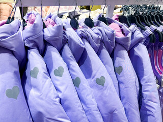  A row of purple jackets with heart patterns hangs neatly on hangers in a store. The jackets feature soft pink linings, displayed uniformly, evoking a sense of warmth and coziness.

