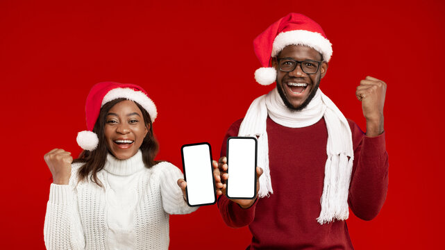 Great Christmas News. African American Couple Showing Phones Empty Screens Wearing Santa Hats And Gesturing Yes Smiling To Camera On Yellow Studio Background. Great Xmas Offer, Mockup