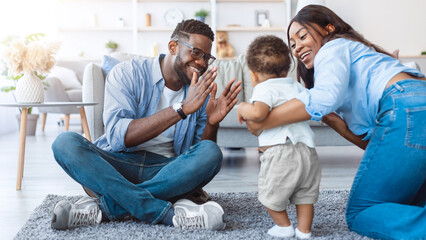 Family Playing Together. Adorable cute baby boy learning how to walk, happy mom proud of little child helping and teaching him sitting on floor carpet, dad clapping hands. First Steps.