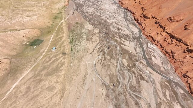 A top-down aerial view of a wide, arid riverbed where faint vehicle tracks follow braided channels, bordered sharply on the right by a massive, eroded red earthen cliff facev