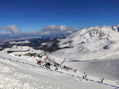 A dramatic high-altitude ski resort with snow-covered slopes and peaks under a brilliant blue sky, showing people near a mountain station.