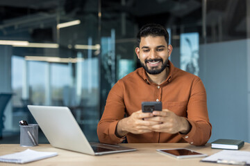 Young indian businessman sitting at his desk in a contemporary office, happily navigating on his smartphone, representing concepts like mobile communication, business technology, and connectivity