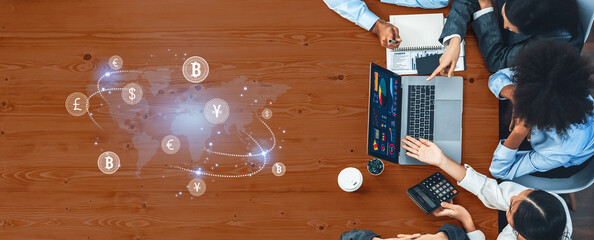 A diverse business team collaborates over a laptop at a wooden table, discussing financial strategies with cryptocurrency icons and global data visualization elements. Trope