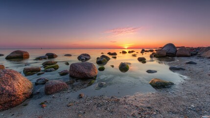 Rocks scatter along a serene beach as the sun sets on the horizon. The sky displays hues of orange and purple while water reflects the tranquil scene. Nature shows its beauty in this peaceful moment.