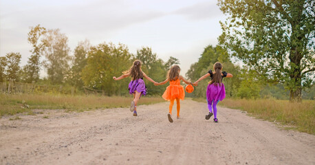 Children dressed up in Halloween costumes are running along a country road with an orange pumpkin-shaped bucket to collect sweets