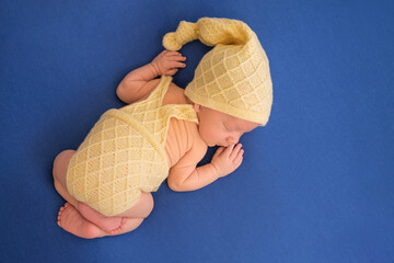 A newborn baby in a knitted yellow suit sleeps sweetly on a blue background. Newborn photo shoot