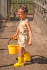 A child in yellow rain boots carries a bucket of water and a bowl of corn to the chickens. The girl helps her grandmother in the village with farming, taking care of poultry