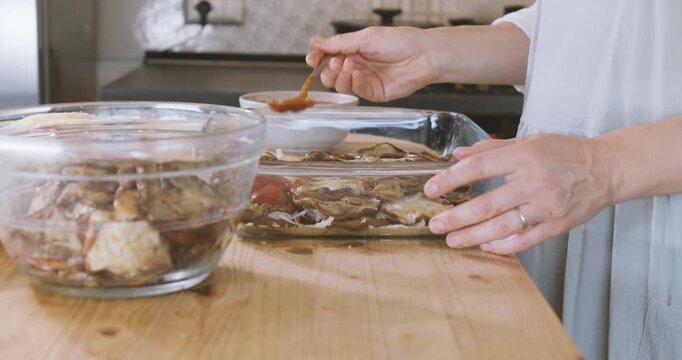 Woman prepares eggplant parmigiana, putting tomato sauce on fried slices of eggplant into lasagna pan