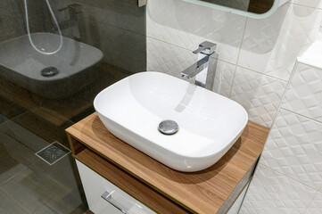 A stylish bathroom features a white oval vessel sink atop a wooden cabinet, paired with a chrome faucet. White textured tiles adorn the wall. Reflected shower visible