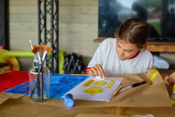 Girl boy focused on painting with yellow paint at art class, sitting at table with brushes, paper and colorful background materials