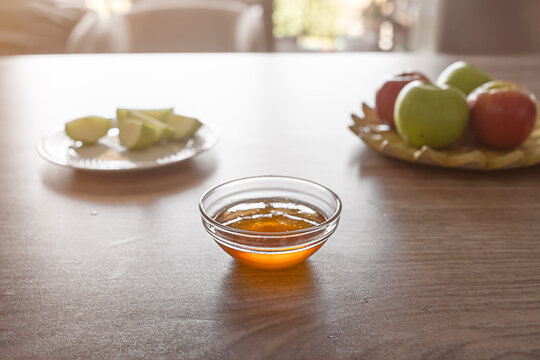 A bowl of honey sits on a wooden table next to fresh apple slices and whole apples. The atmosphere is warm and inviting, perfect for Rosh Hashanah festivities. - Powered by Adobe