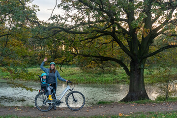 Happy mother with child in safety helmet cycling together on bike with child seat in autumn park under large tree. Concept of family activities and healthy outdoor lifestyle with children.