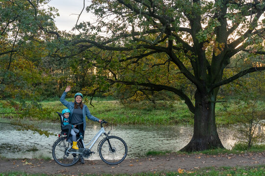 Happy mother with child in safety helmet cycling together on bike with child seat in autumn park under large tree. Concept of family activities and healthy outdoor lifestyle with children. - Powered by Adobe