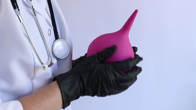 A female doctor in a white coat and with a stethoscope holds a large enema in her hands, close-up. A female doctor squeezes a pink rubber enema, checking for serviceability. Medical item. Nurse