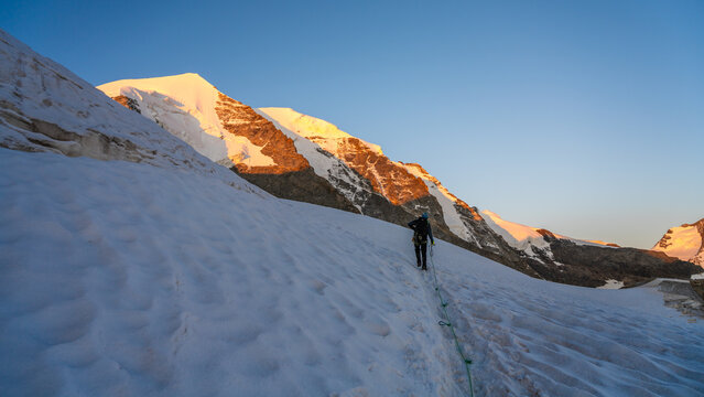 A climber ascends the snowy slopes of Piz Palu in the Bernina Range during a summer morning. The bright sun illuminates the majestic peaks and the glacier landscape surrounding the climber.