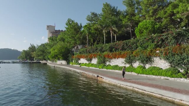 Scenic Promenade: A person walks along a pathway, overlooking a serene body of water, with lush greenery. a castle graces the horizon.