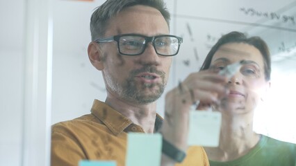 Professional team members brainstorming strategy, writing notes on transparent whiteboard with colorful sticky papers during collaborative meeting in office. Business people at work