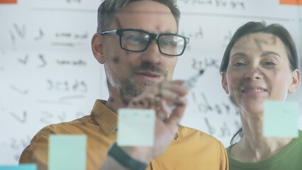 Professional team members brainstorming strategy, writing notes on transparent whiteboard with colorful sticky papers during collaborative meeting. Business people concept