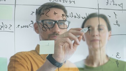 Business team of professionals brainstorming near glass board, using sticky notes for strategic planning, writing calculations, sharing innovative ideas during collaborative work