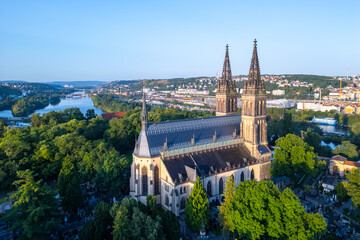Fototapeta premium Golden sunlight bathes the Basilica of St. Peter and St. Paul in Prague during a tranquil morning. Surrounded by lush greenery, the historic structure stands majestically against the skyline.