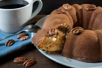 Vanilla donut with nuts, cut on a white plate, a white cup with black coffee, a spoon and a blue napkin on a table, angle view