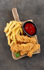 Crispy chicken strips with ketchup and French fries on a wooden board. Fast food. Gray background