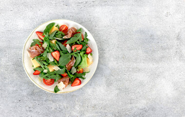 Italian arugula salad with prosciutto, strawberries, melon, cheese on gray background. Top view. Copy space.