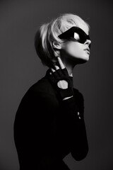 Stylish young woman in dark attire, wearing oversized sunglasses, striking a pose with a confident expression against a minimalist black background.