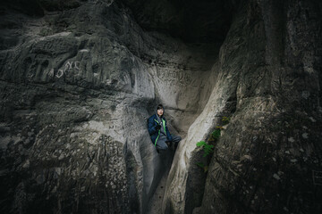 A boy in a hooded jacket sits in a narrow rock crevice covered with carvings and graffiti in a shadowy forest canyon.