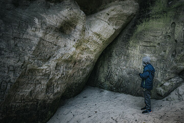 A boy in a hooded jacket sits in a narrow rock crevice covered with carvings and graffiti in a...