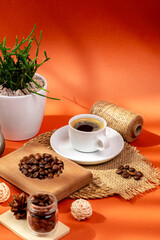 Cup of hot tasty coffee on wooden desk, coffee beans, candle and house plant in a beautiful cozy composition from above on orange background in sunlight. Americano, espresso and roasted coffee grain