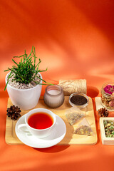 Mug of hot tasty tea on wooden desk, herbal tea leaves in glass bowls, candle and house plant in a beautiful cozy composition from above on orange background in sun light.