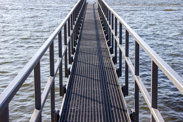 Obraz premium Metal walkway extends over calm water, leading to distant horizon, surrounded by gentle waves and reflections, creating a serene and inviting atmosphere for exploration