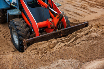 Close up of a red and blue front loader leveling sand with its metal blade. Rugged tires and...