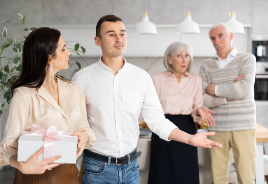 Positive young man and woman holding present going away while old parents watching them indignantly standing in their back