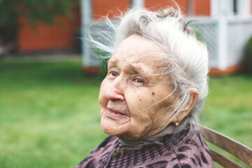 Elderly woman with gray hair sitting outdoors on a bench, looking thoughtfully into the distance, concept of aging and memories