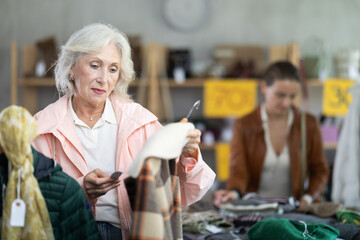 Elderly woman with gray hair stands near the hangers and chooses jackets and a woolen coat in front of a customer who chooses a sweater. Buying clothes and sweaters for autumn and winter
